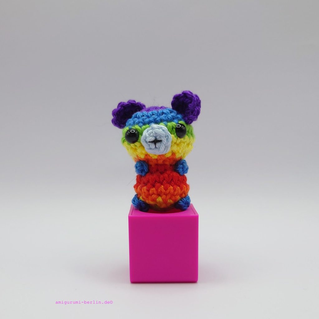 ein regenbogenfarbener gehäkelter Bär auf einem pinkfarbenen Block - a rainbow-coloured crocheted bear on a pink block