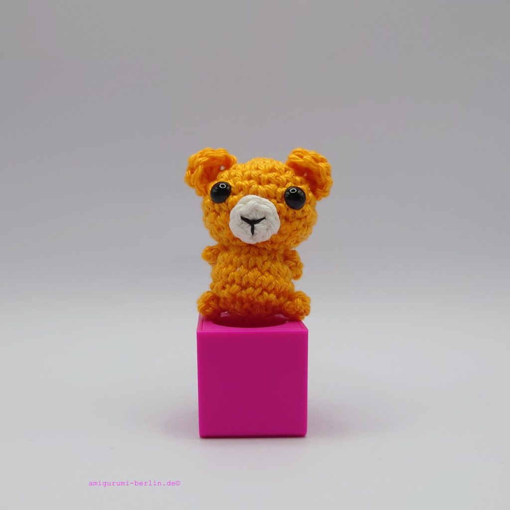 ein mangofarbener gehäkelter Bär auf einem pinkfarbenen Block a mango-coloured crocheted bear on a pink block