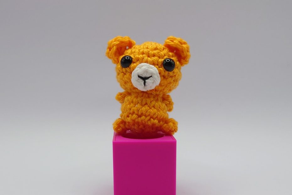 ein mangofarbener gehäkelter Bär auf einem pinkfarbenen Block a mango-coloured crocheted bear on a pink block