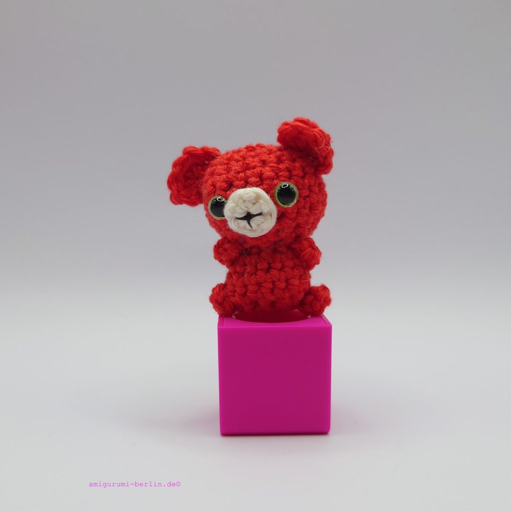 ein signalroter gehäkelter Bär mit grünen Augen auf einem pinkfarbenen Block a signal red crocheted bear with green eyes on a pink block