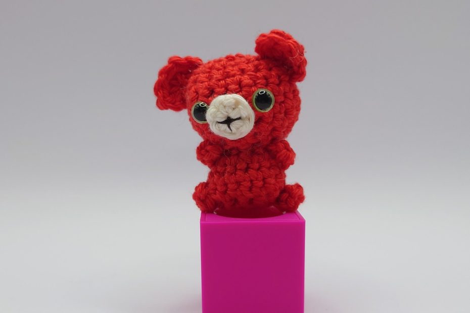ein signalroter gehäkelter Bär mit grünen Augen auf einem pinkfarbenen Block a signal red crocheted bear with green eyes on a pink block