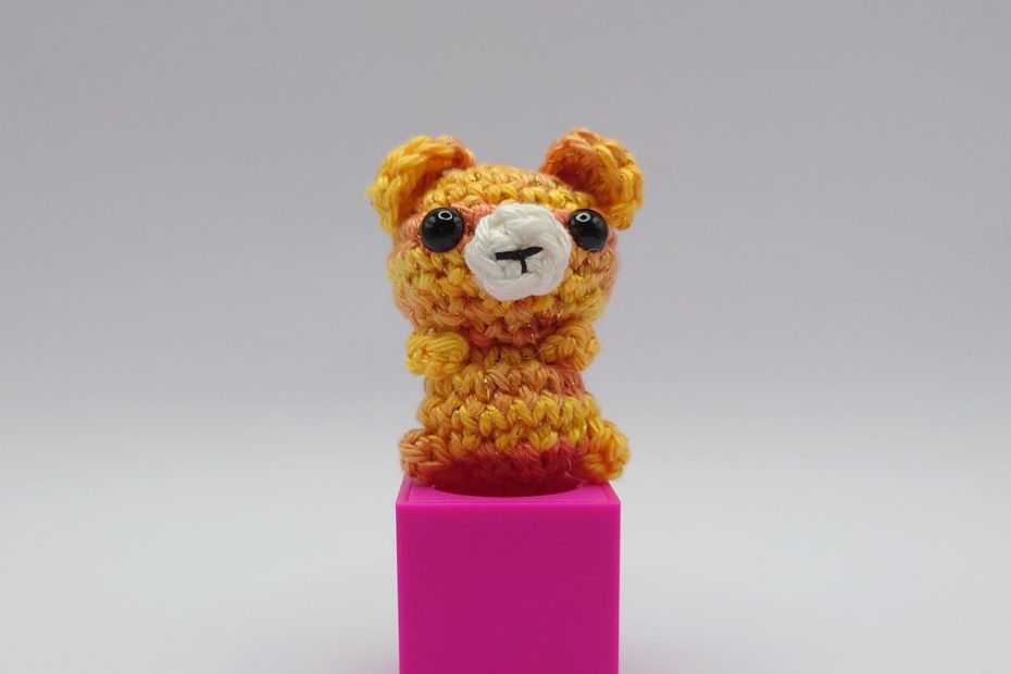 ein gold orangefarbener gehäkelter Bär auf einem pinkfarbenen Block a golden orange crocheted bear on a pink block