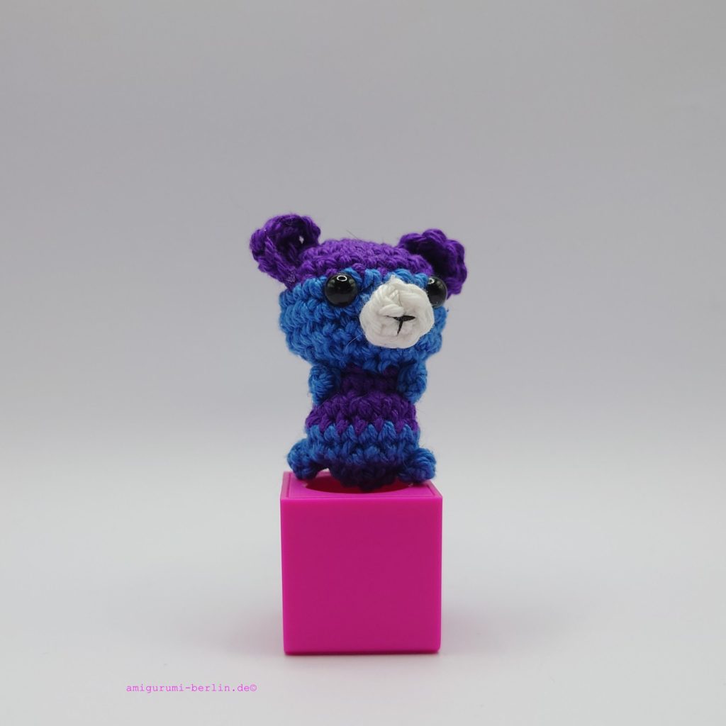 ein violetter und marineblauer gehäkelter Bär auf einem pinkfarbenen Block a purple and navy blue-coloured crocheted bear on a pink block