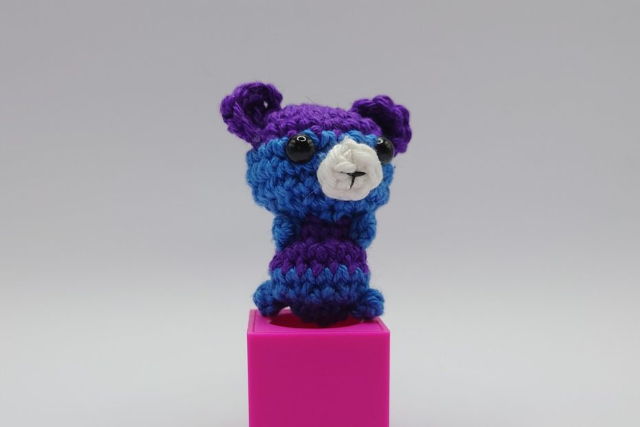 ein violetter und marineblauer gehäkelter Bär auf einem pinkfarbenen Block a purple and navy blue-coloured crocheted bear on a pink block